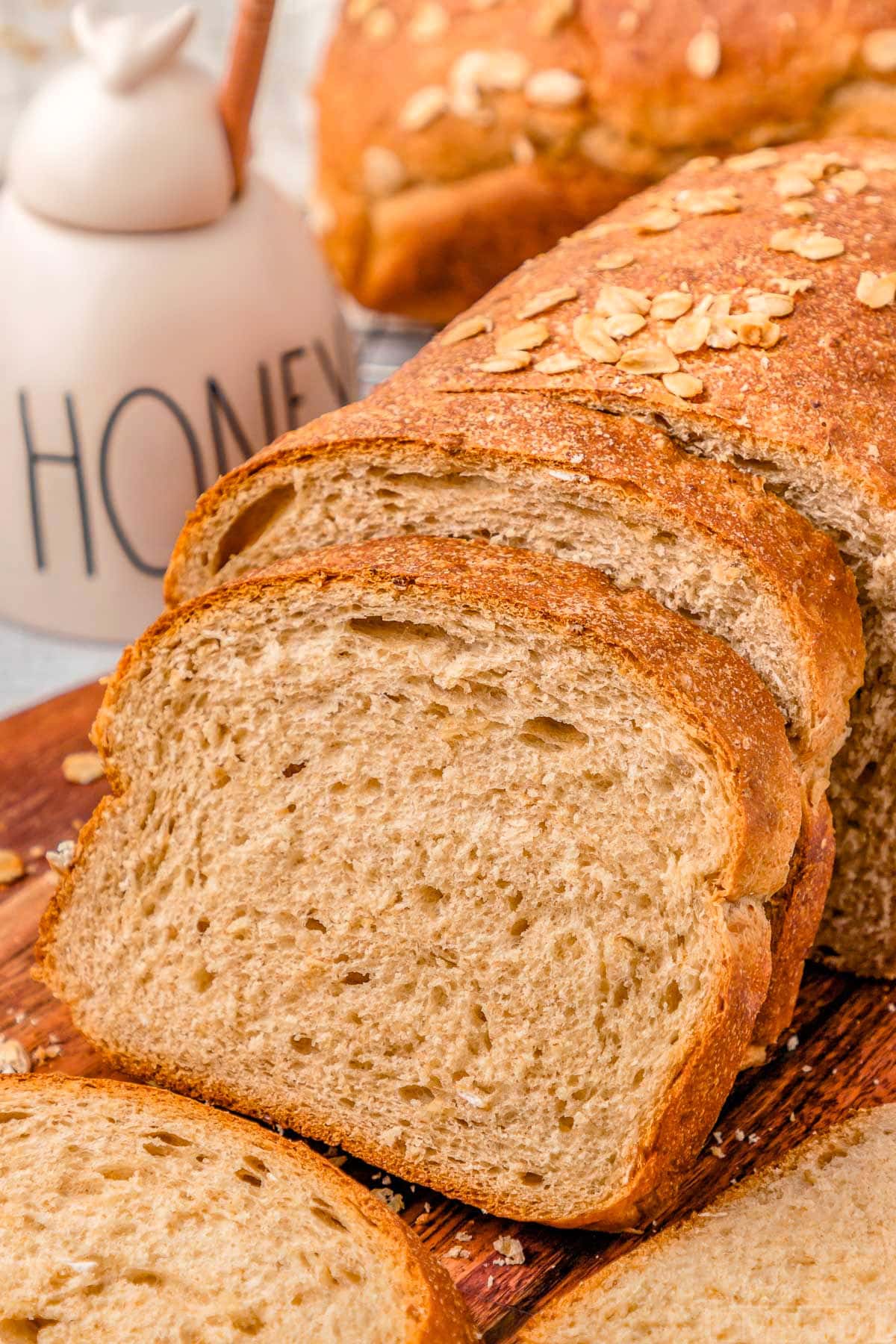 two slices cut off a loaf of wheat bread on a wood board. more bread can be seen in the background next to a white honey jar.