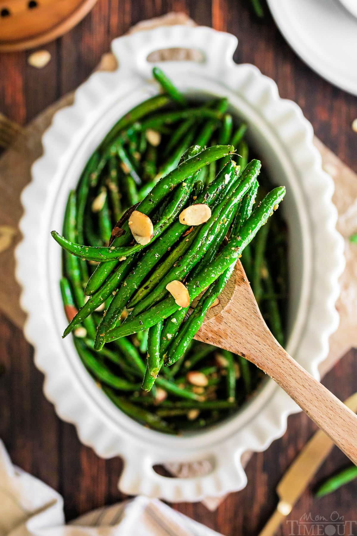 wooden spoon lifting out a serving of garlic green beans out of an oval white serving dish.