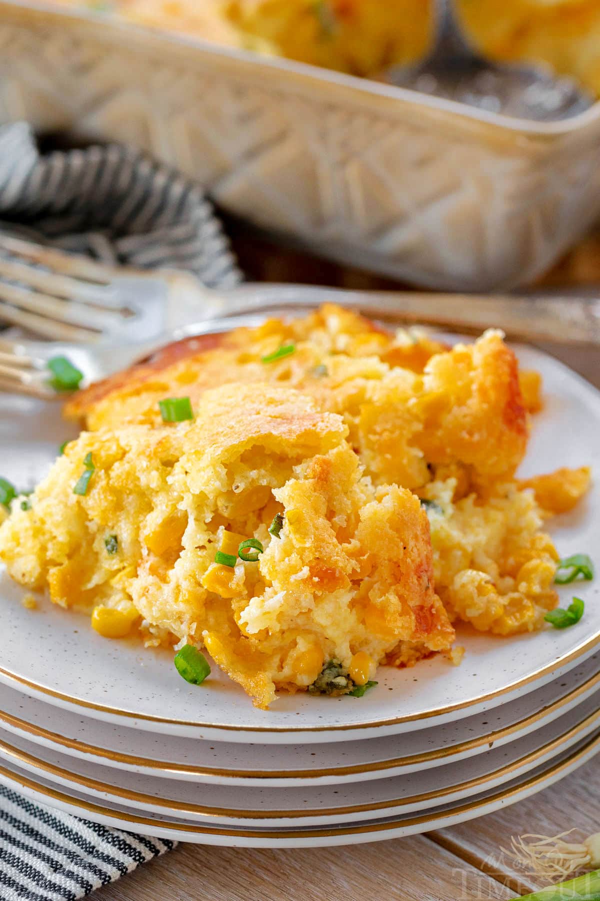 scoop of corn casserole on a plate that is stacked on three more plates. green onions have been sprinkled over the top. the casserole dish can be seen in the background.