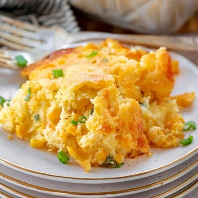 scoop of corn casserole on a plate that is stacked on three more plates. green onions have been sprinkled over the top. the casserole dish can be seen in the background.