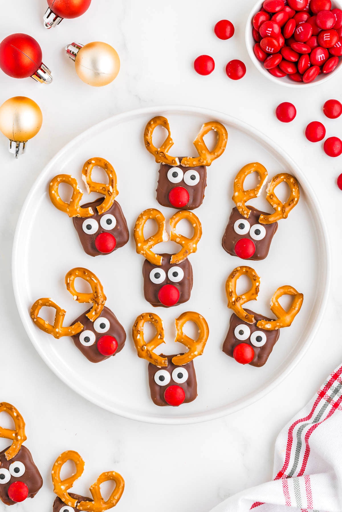 small white plate with seven pretzel reindeer on it surrounded with more treats, candies and ornaments.