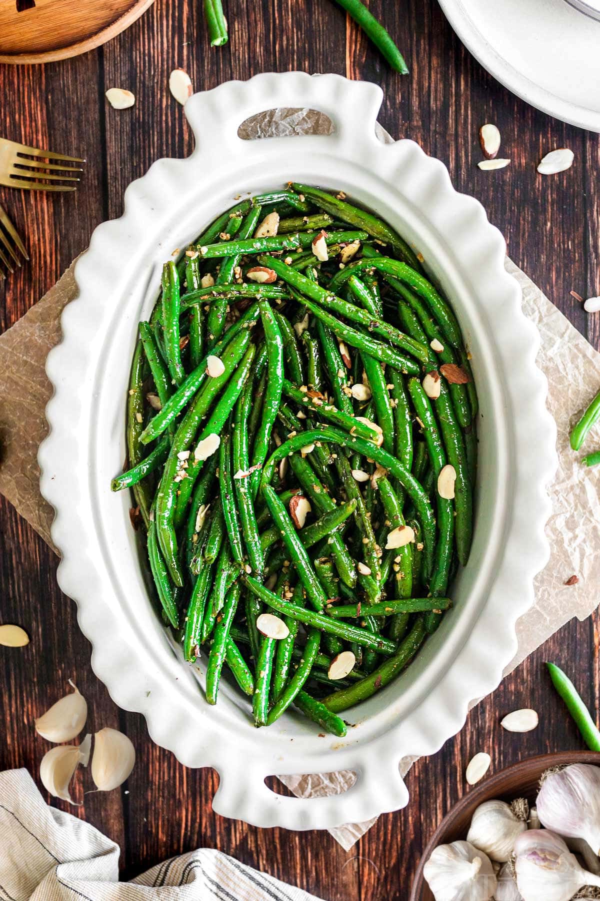 top down view of large white oval serving bowl filled with bright green garlic green beans. bowl is on a dark wood surface.