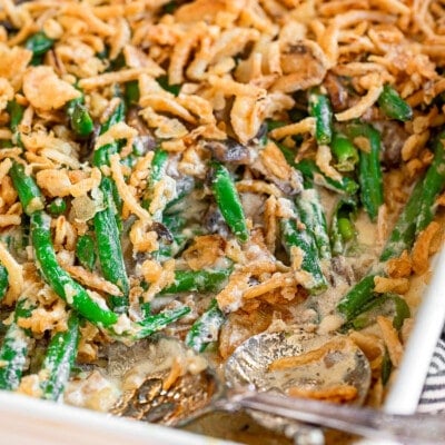 green bean casserole in a large white baking dish topped with fried onions. two silver serving spoons are in the dish with a serving or two removed.