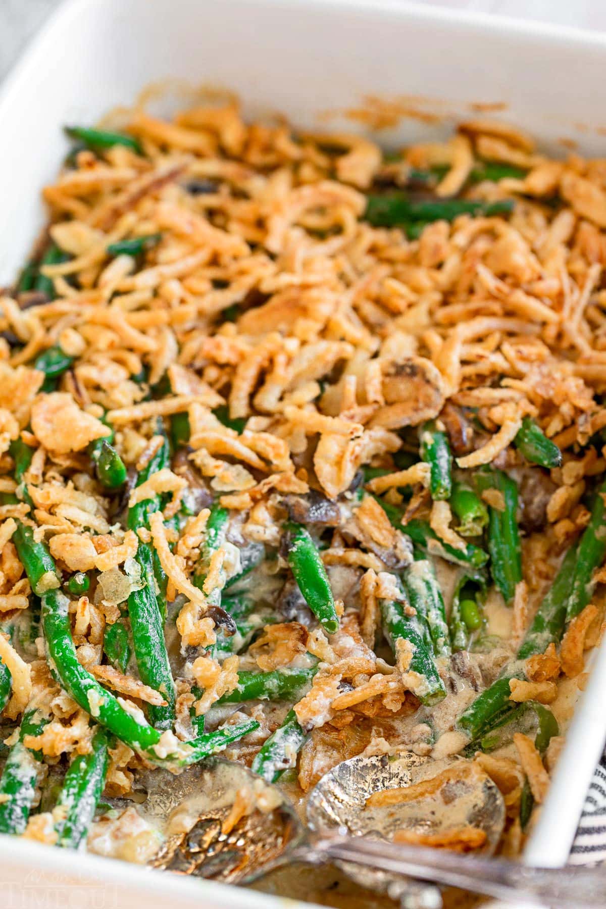 green bean casserole in a large white baking dish topped with fried onions. two silver serving spoons are in the dish with a serving or two removed.
