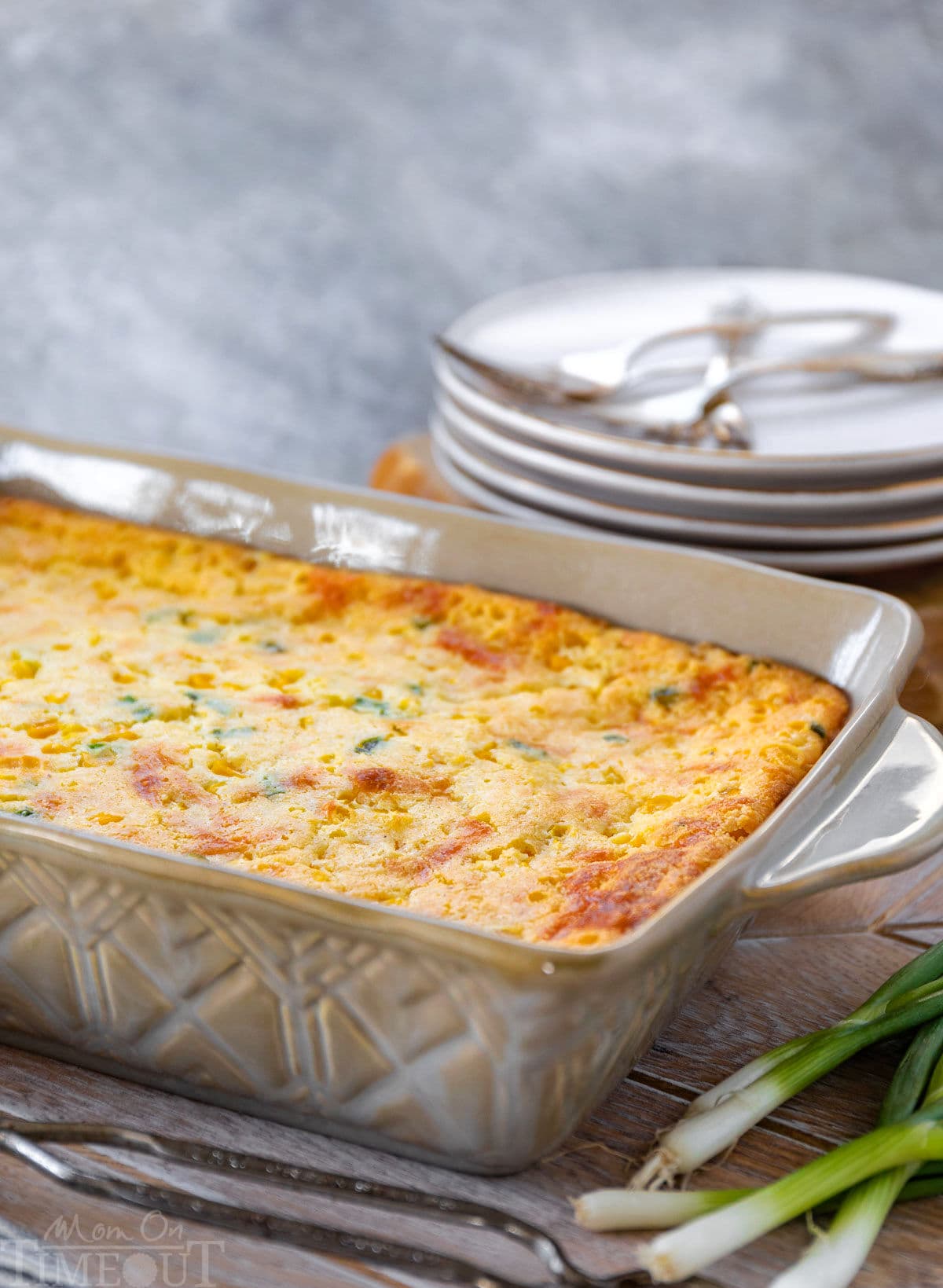 corn casserole recipe in a brown baking dish ready to be served. stack of plates behind the casserole and green onions in the front.