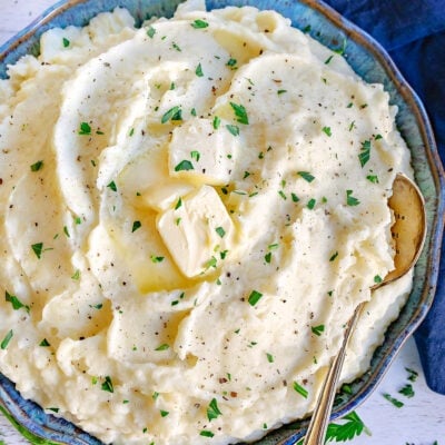 top down look at a blue and green bowl filled with fluffy mashed potatoes. slices of butter, pepper and chopped parsley garnish the mashed potatoes.