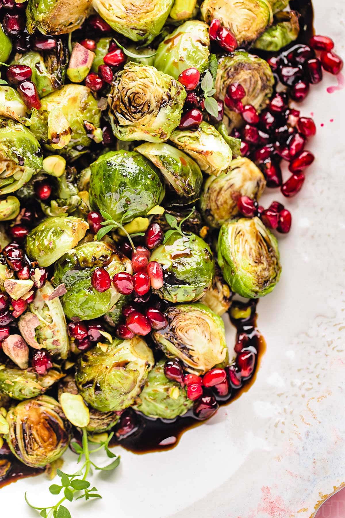 Top down view of roasted brussels sprouts on a pink and white serving dish. the sprouts are topped with pomegranate seeds and thyme along with a balsamic glaze.