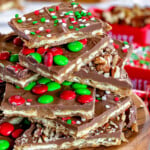Square pieces of Christmas Crack cut and stacked on a round wood board. The toffee is topped with candy and sprinkles.