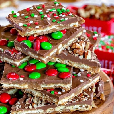 Square pieces of Christmas Crack cut and stacked on a round wood board. The toffee is topped with candy and sprinkles.