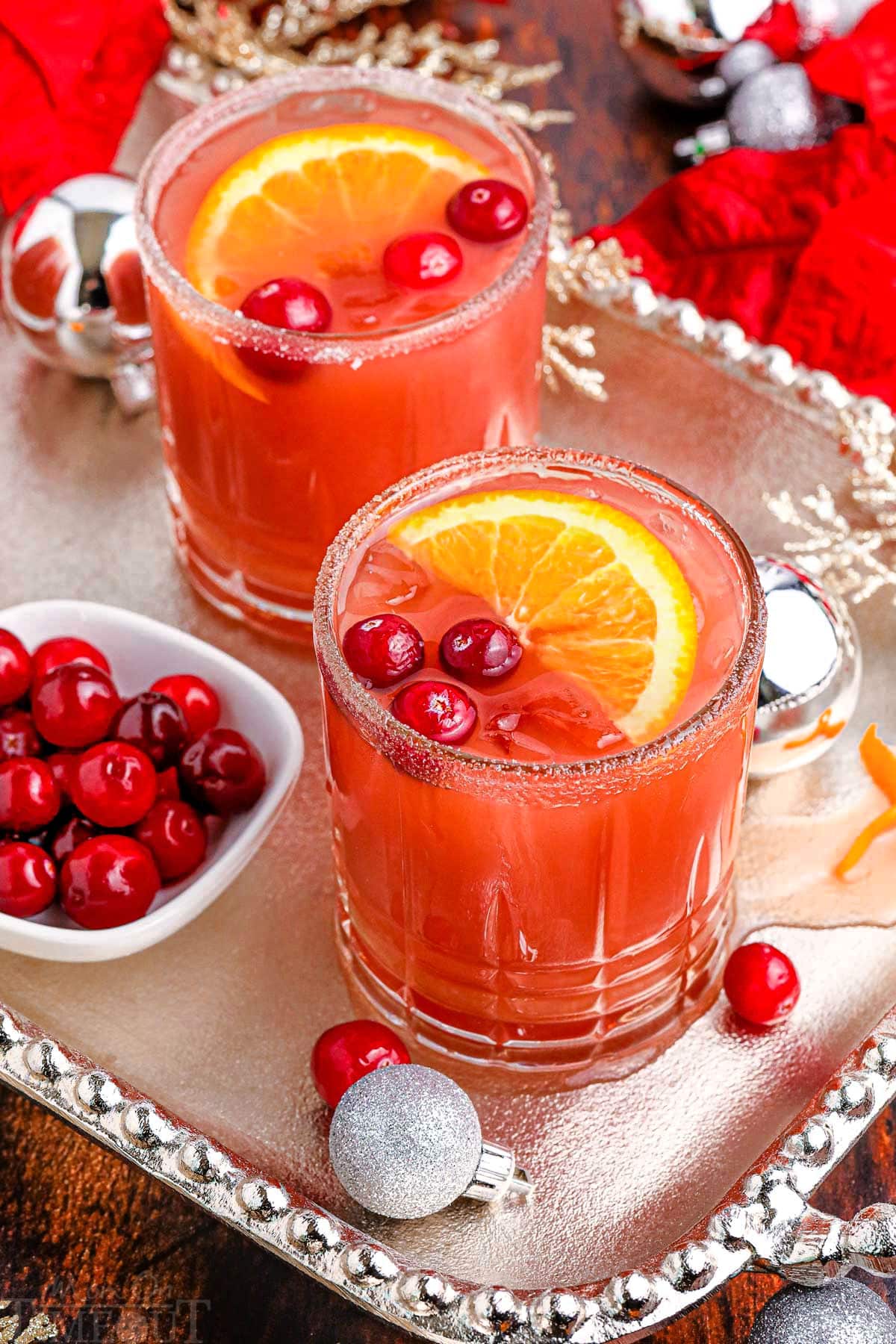 two christmas cranberry cocktails on a silver serving platter garnished with fresh cranberries and orange slices.