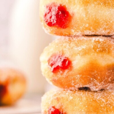 Three jelly donuts stacked on a small white plate. The bright red jelly can be seen on the sides of the sugar coated donuts.