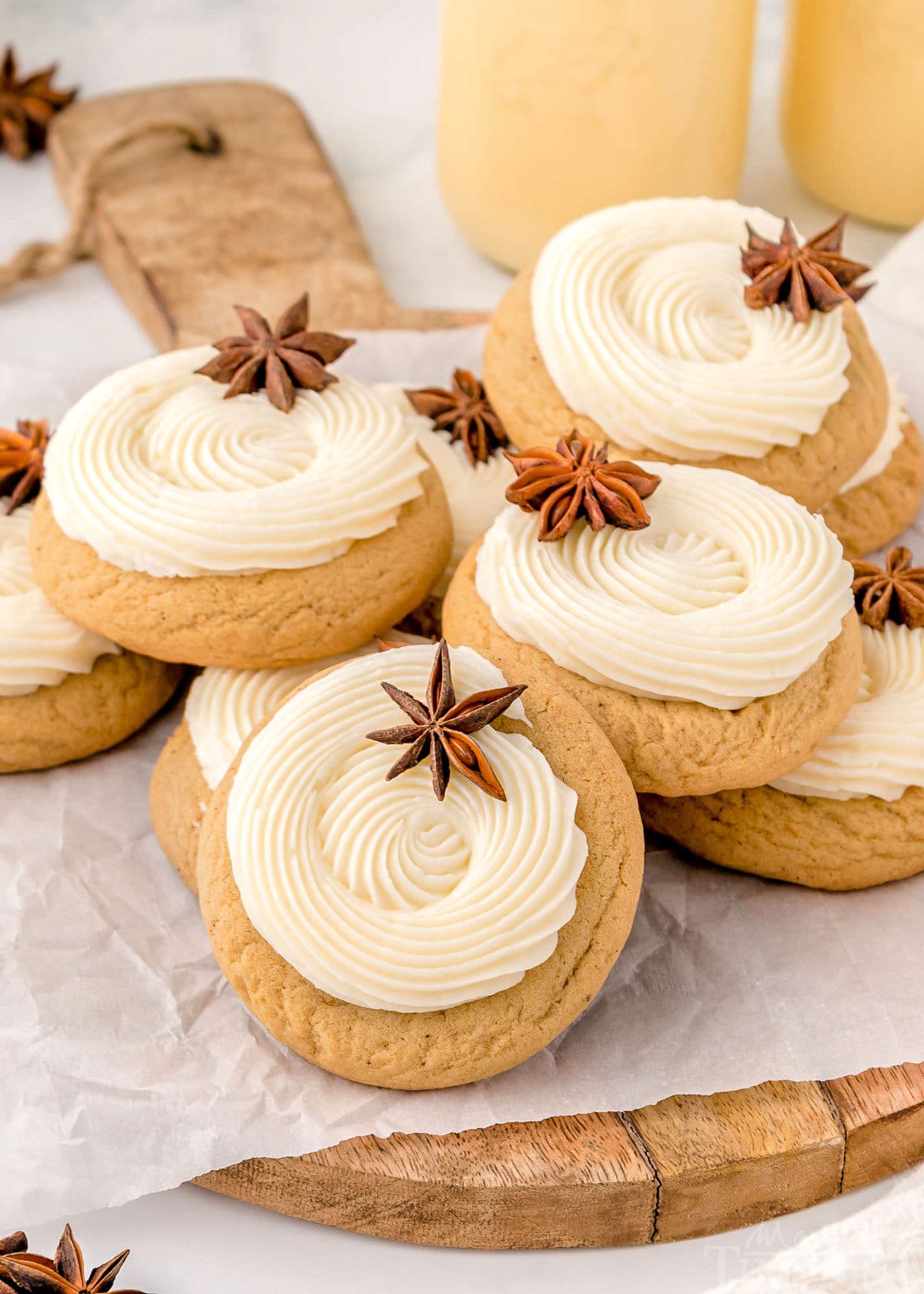 pile of eggnog cookies on wood cutting board. cookies have been frosted with eggnog frosting in a thick swirl and are garnished with anise. two cups of eggnog can be seen in the background.