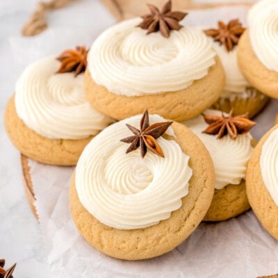 pile of eggnog cookies on wood cutting board. cookies have been frosted with eggnog frosting in a thick swirl and are garnished with anise.