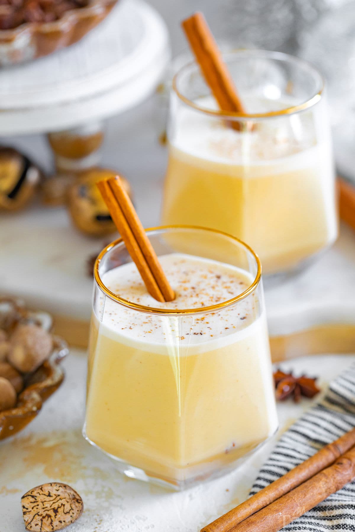 two glasses of eggnog garnished with freshly grated nutmeg and a cinnamon stick. more cinnamon sticks, whole nutmeg and star anise are scattered about the two glasses.