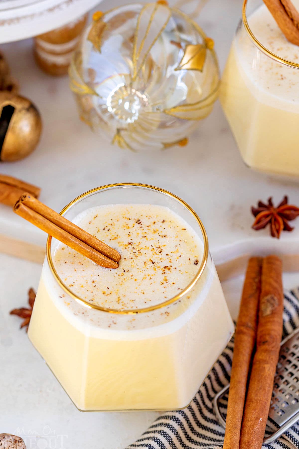gold rimmed short glass filled with homemade eggnog with freshly grated nutmeg on top and a cinnamon stick as a garnish. another glass can be seen in the background.