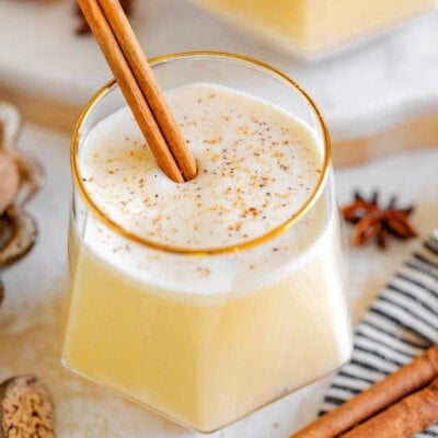 gold rimmed short glass filled with homemade eggnog with freshly grated nutmeg on top and a cinnamon stick as a garnish. another glass can be seen in the background.