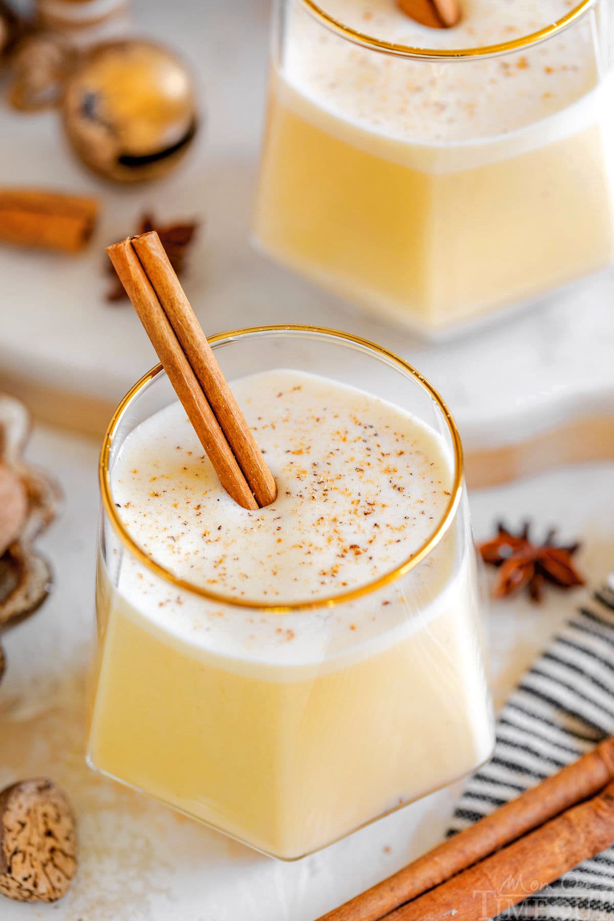 gold rimmed short glass filled with homemade eggnog with freshly grated nutmeg on top and a cinnamon stick as a garnish. another glass can be seen in the background.