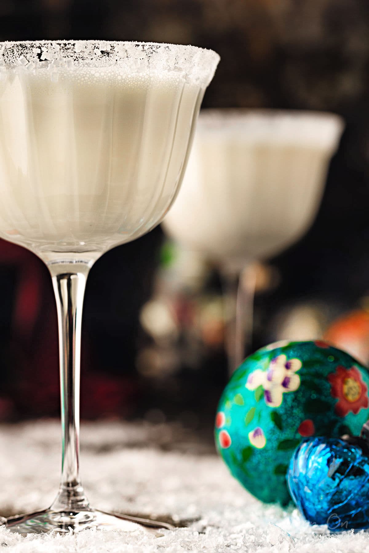 two white martinis in tall glass dusted with powdered sugar for a snowy look. the cocktails is sitting next to vintage ornaments. bottles of vodka and white chocolate liqueur can be seen in the background.