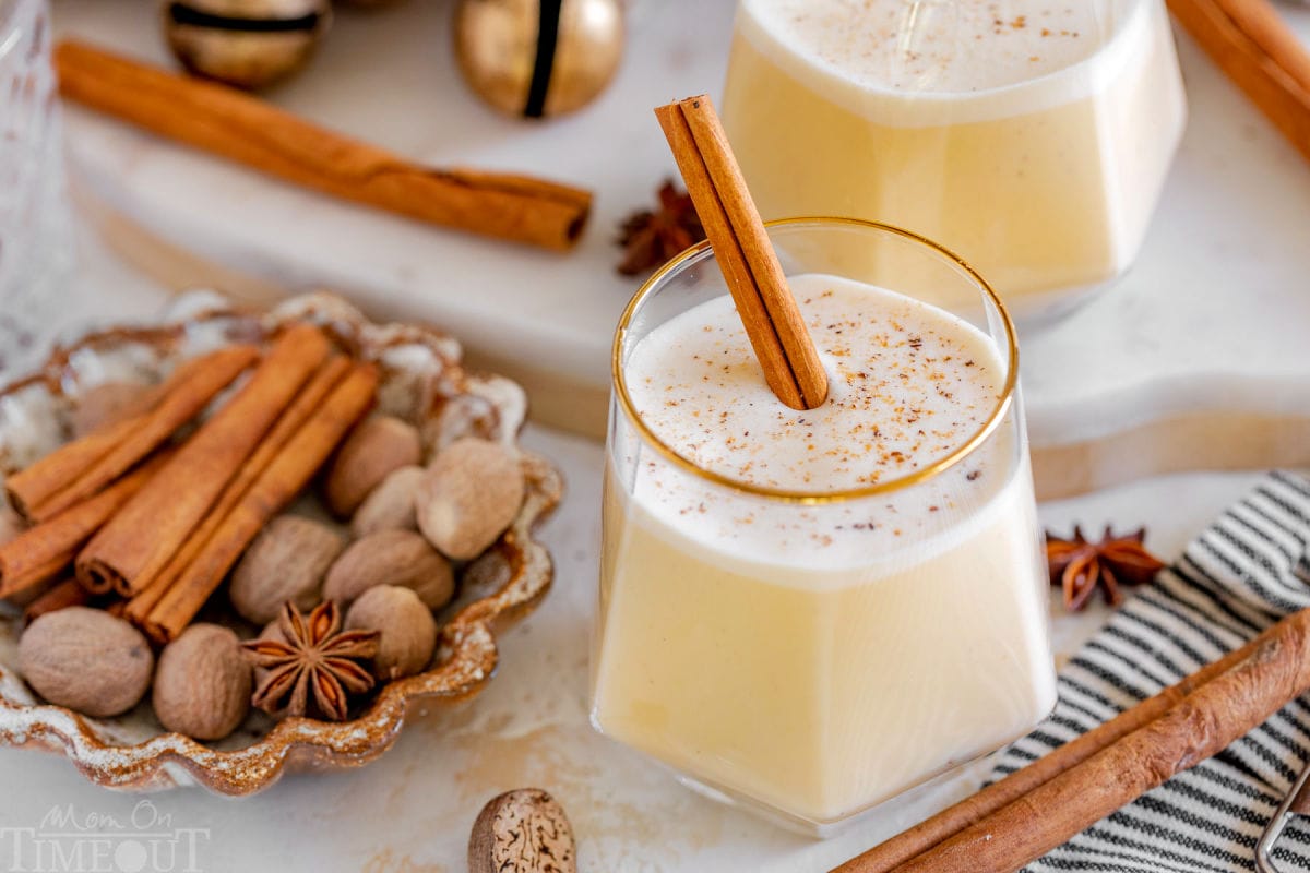 wide shot of two glasses of eggnog garnished with freshly grated nutmeg and a cinnamon stick. more cinnamon sticks, whole nutmeg and star anise are scattered about the two glasses.
