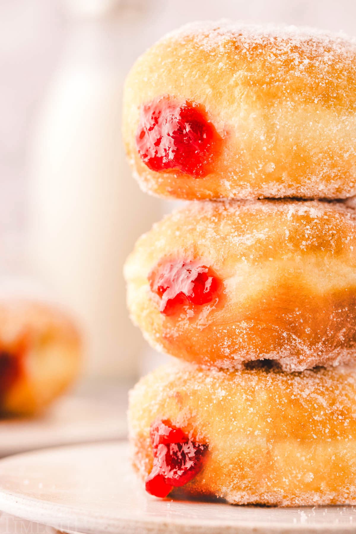 Three jelly donuts stacked on a small white plate. The bright red jelly can be seen on the sides of the sugar coated donuts.