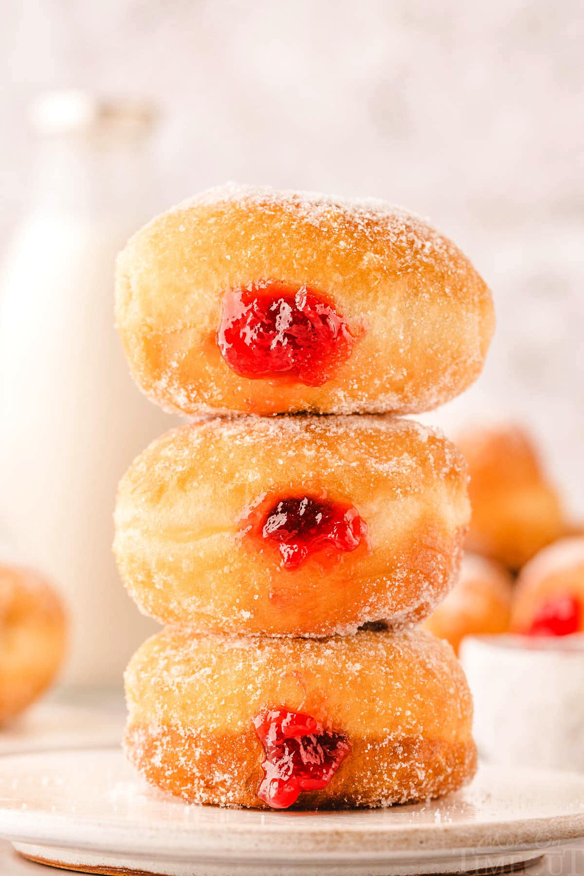 stack of three jelly donuts filled with raspberry jam. more donuts can be seen in the background.