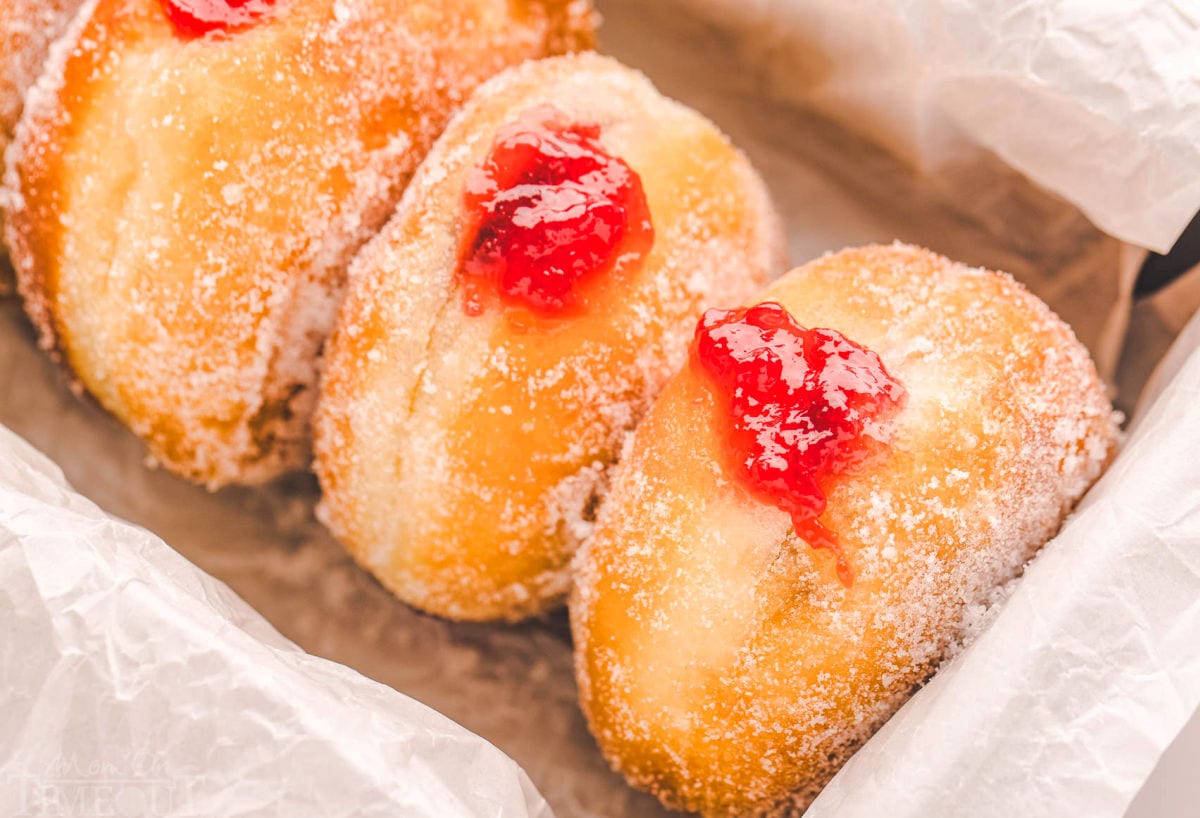 four sufganiyot donuts in a parchment lined container. the jelly is oozing out of the donuts.