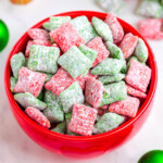 red bowl filled with red and green puppy chow. the bowl is on a white surface which christmas decorations in the background.