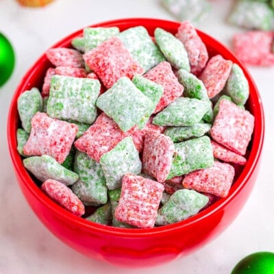 red bowl filled with red and green puppy chow. the bowl is on a white surface which christmas decorations in the background.