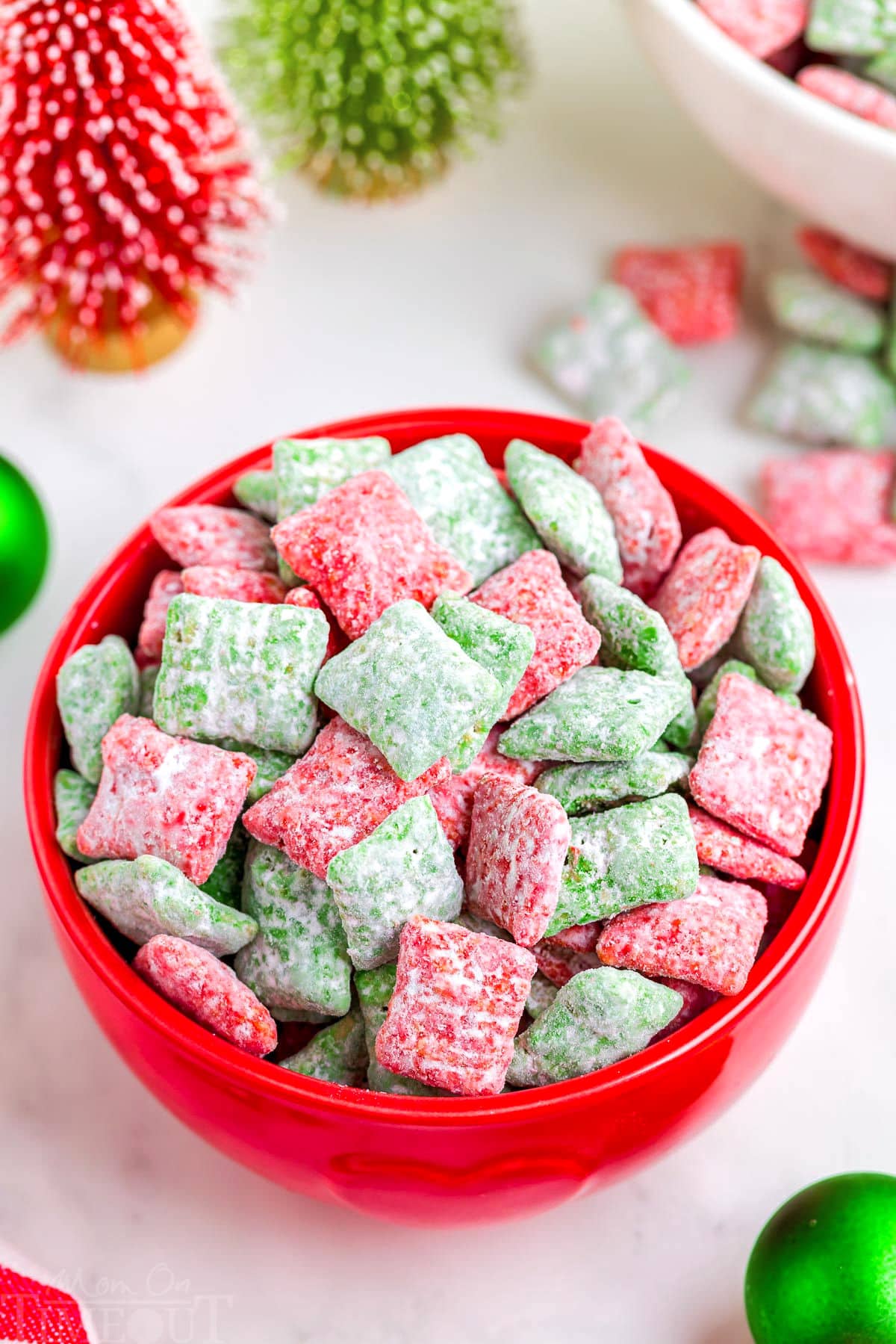 red bowl filled with red and green puppy chow. the bowl is on a white surface which christmas decorations in the background.