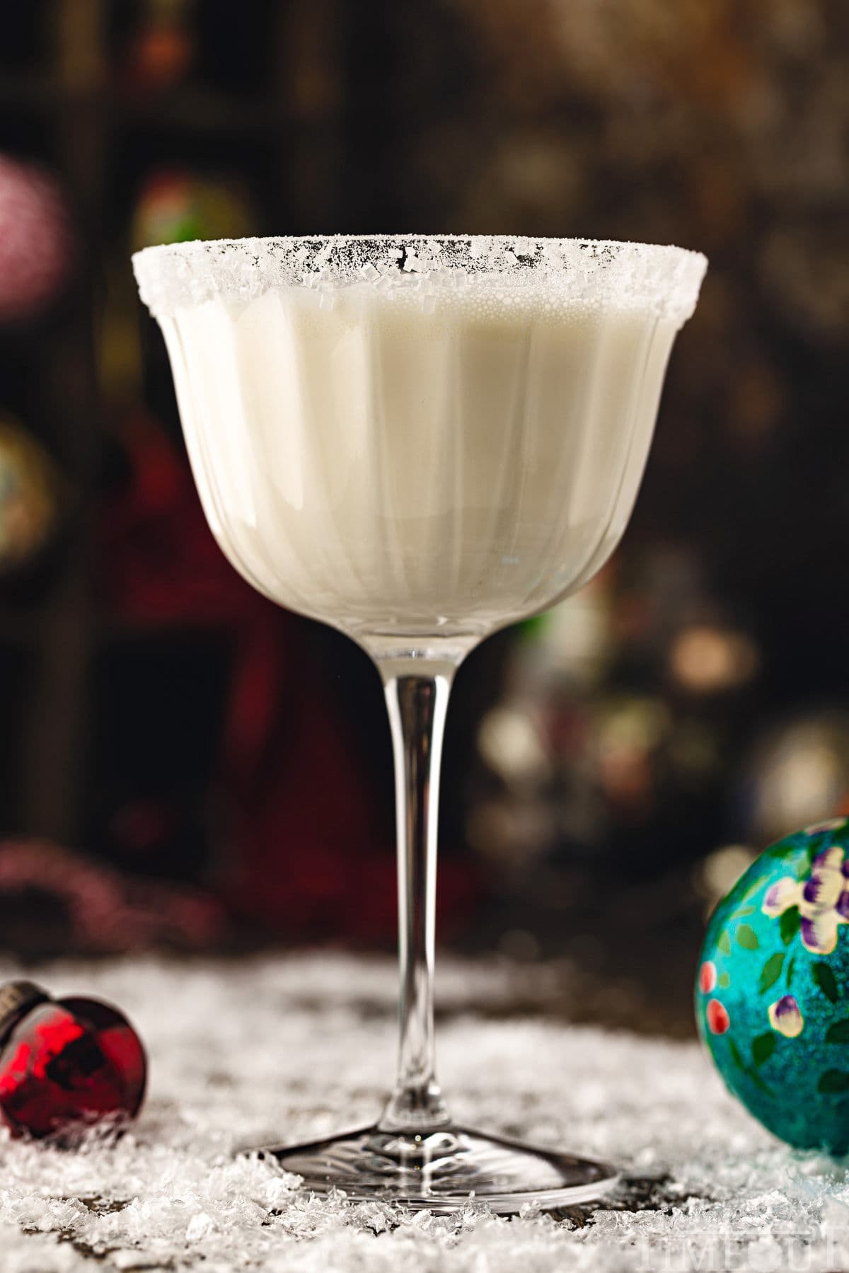 white chocolate martini cocktail in tall glass dusted with powdered sugar for a snowy look. the cocktails is sitting on a snowy surface with glass ornaments scattered about.