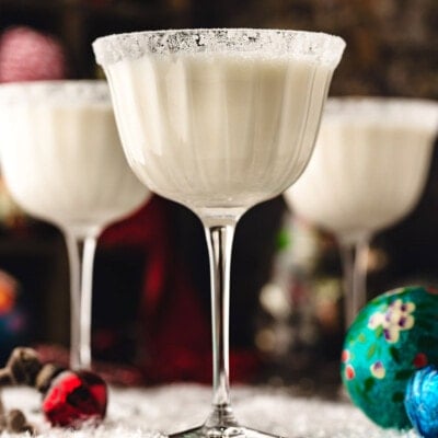 three white chocolate martinis in tall glasses dusted with powdered sugar for a snowy look. the cocktails are sitting on a snowy surface with glass ornaments scattered about.