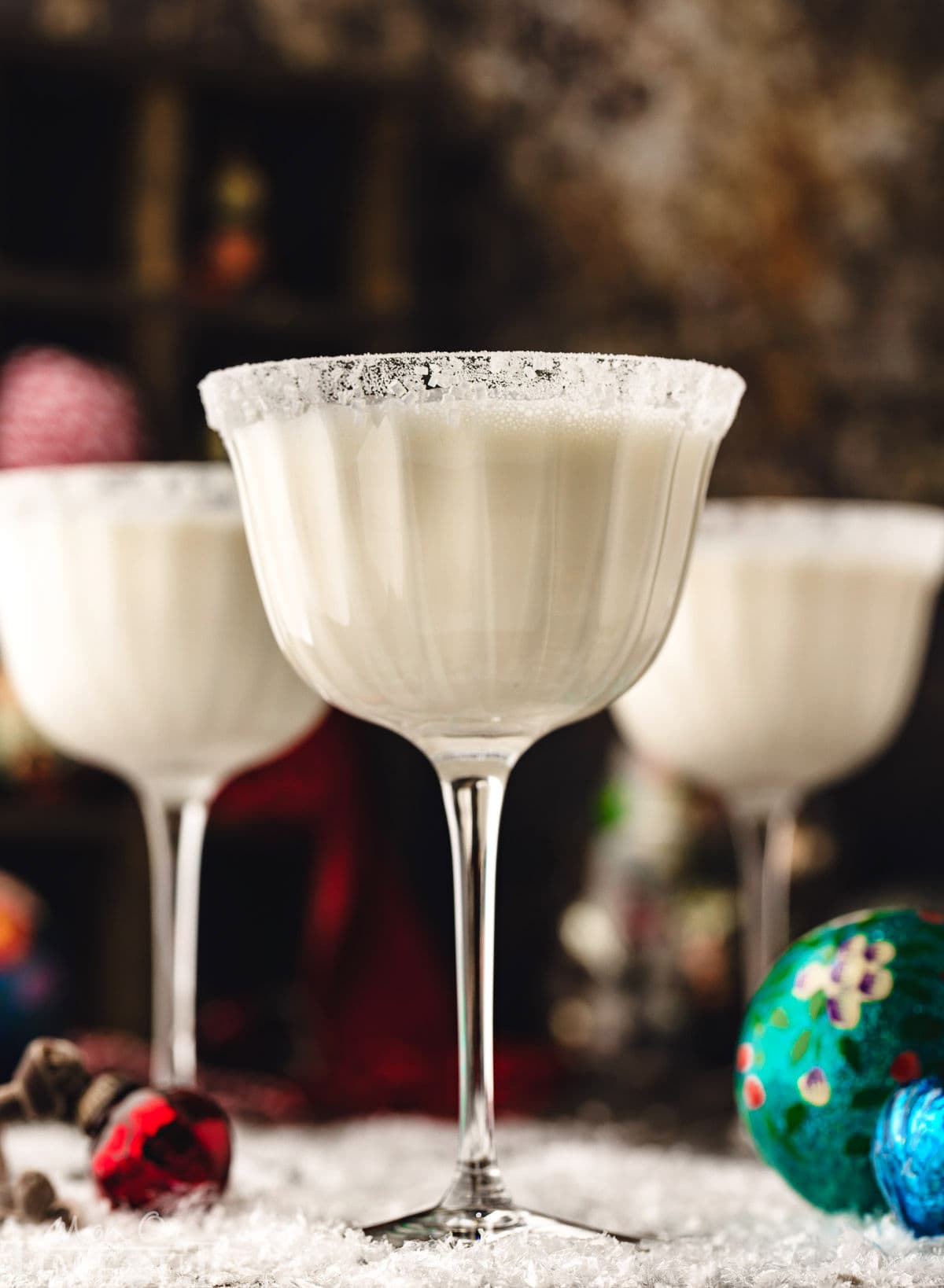 three white chocolate martinis in tall glasses dusted with powdered sugar for a snowy look. the cocktails are sitting on a snowy surface with glass ornaments scattered about.