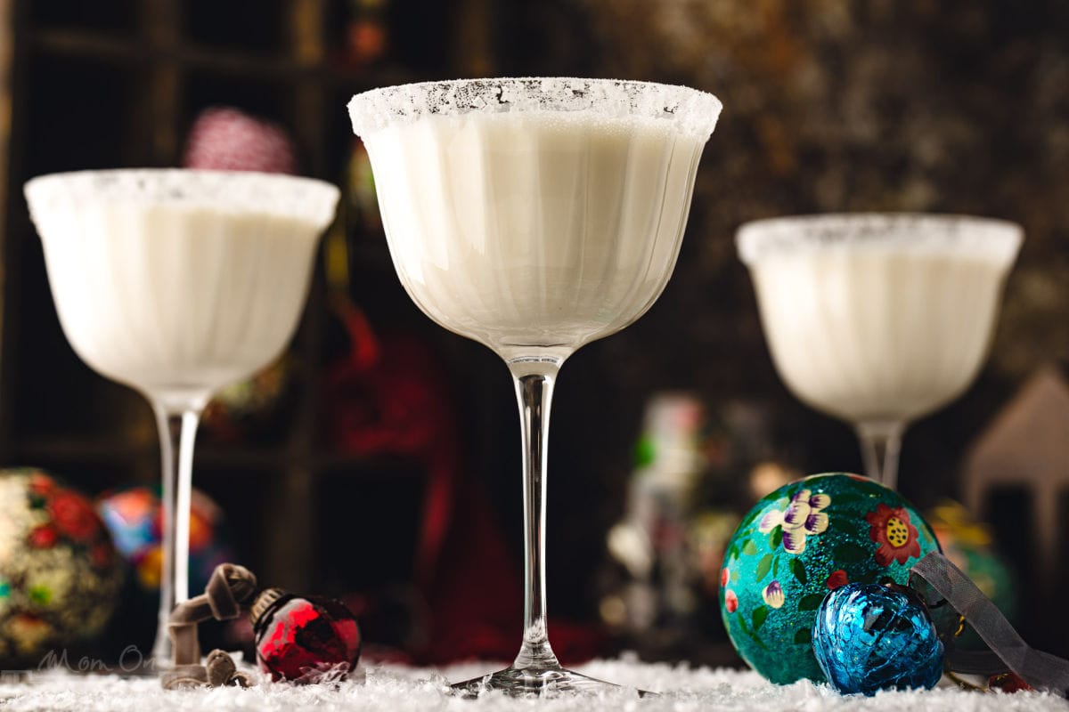 three white chocolate martinis in tall glasses dusted with powdered sugar for a snowy look. the cocktails are sitting on a snowy surface with glass ornaments scattered about.