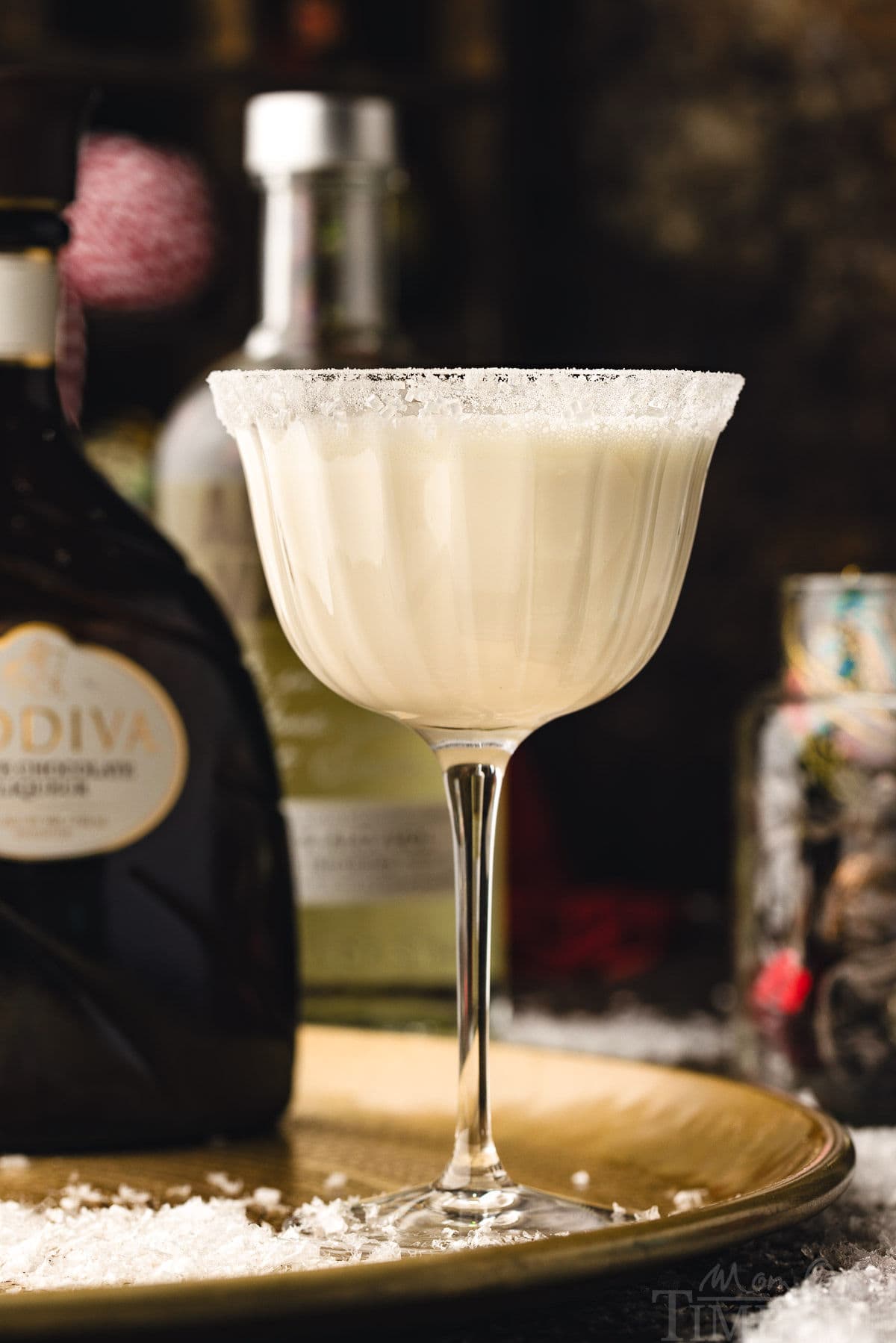 white chocolate martini cocktail in tall glass dusted with powdered sugar for a snowy look. the cocktails is sitting on a gold tray. bottles of vodka and white chocolate liqueur can be seen in the background.