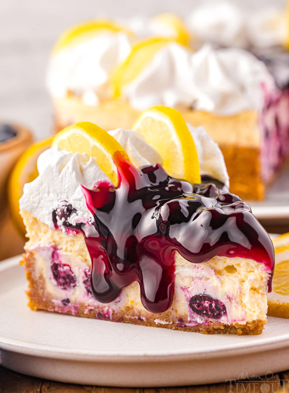 side view of lemon blueberry cheesecake topped with blueberry pie filling and fresh whipped cream. the rest of the cheesecake can be seen in the background.