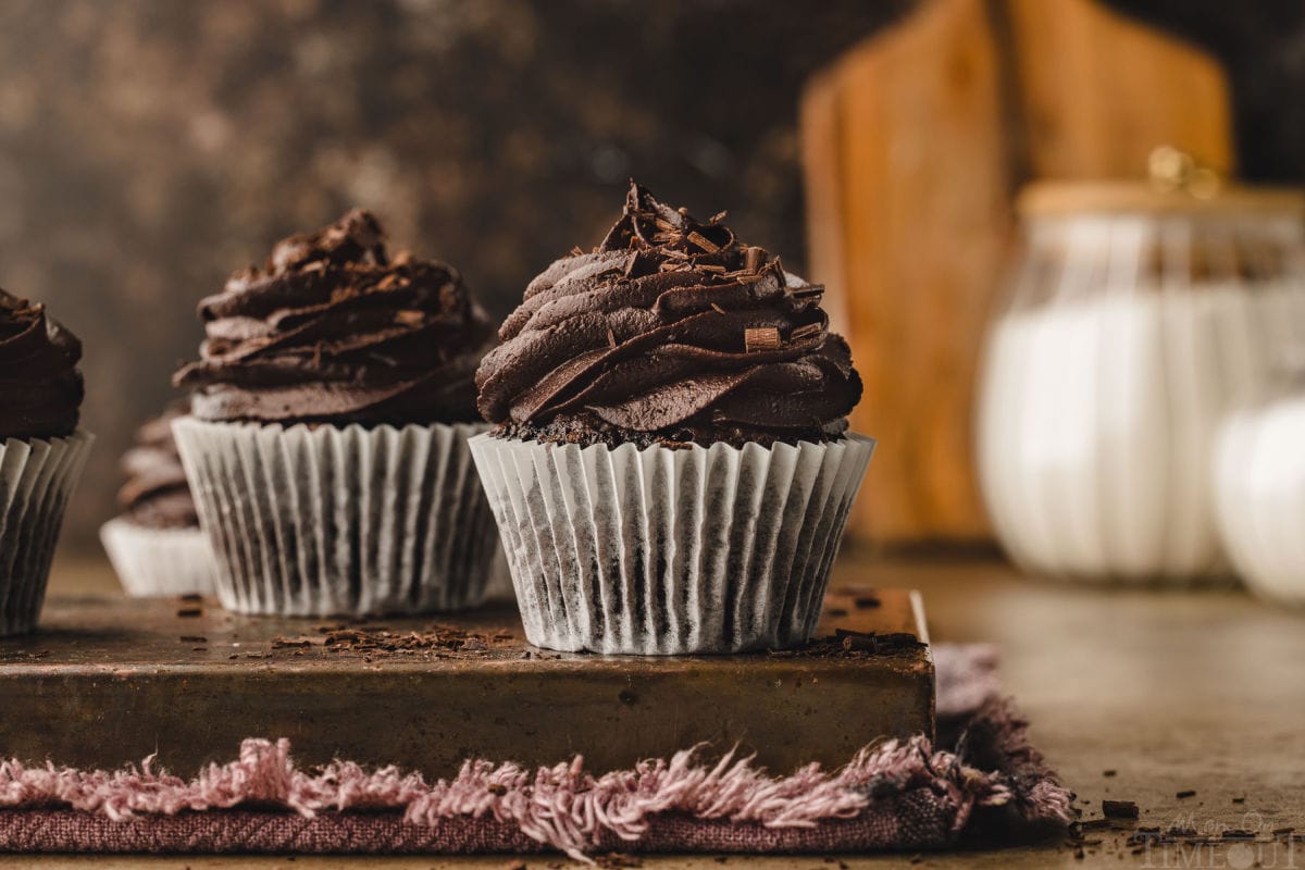 two chocolate cupcakes in white liner topped with chocolate buttercream. cutting board can be seen in the background.