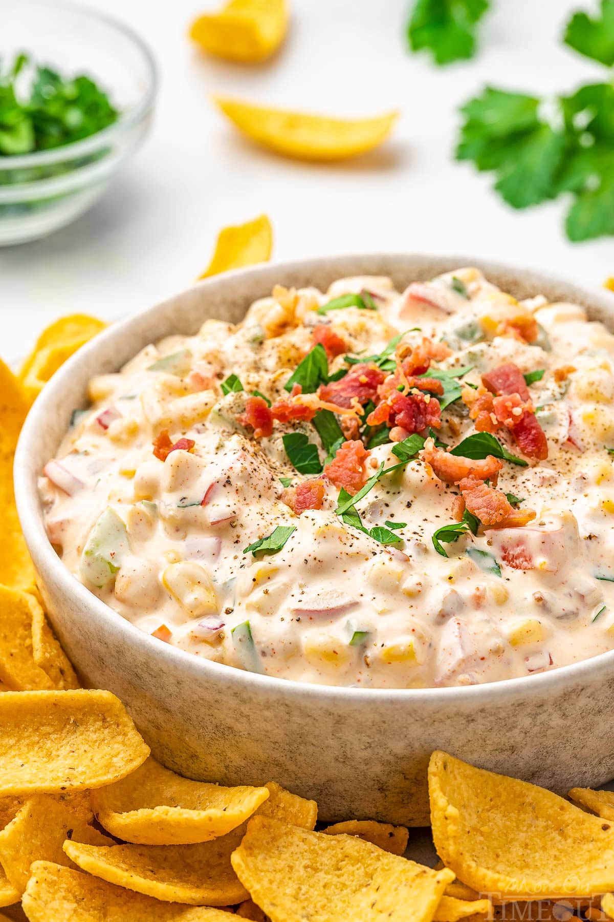 Delicious corn dip recipe in round white bowl topped with bacon bits in parsley surround by fritos scoops ready to be enjoyed.