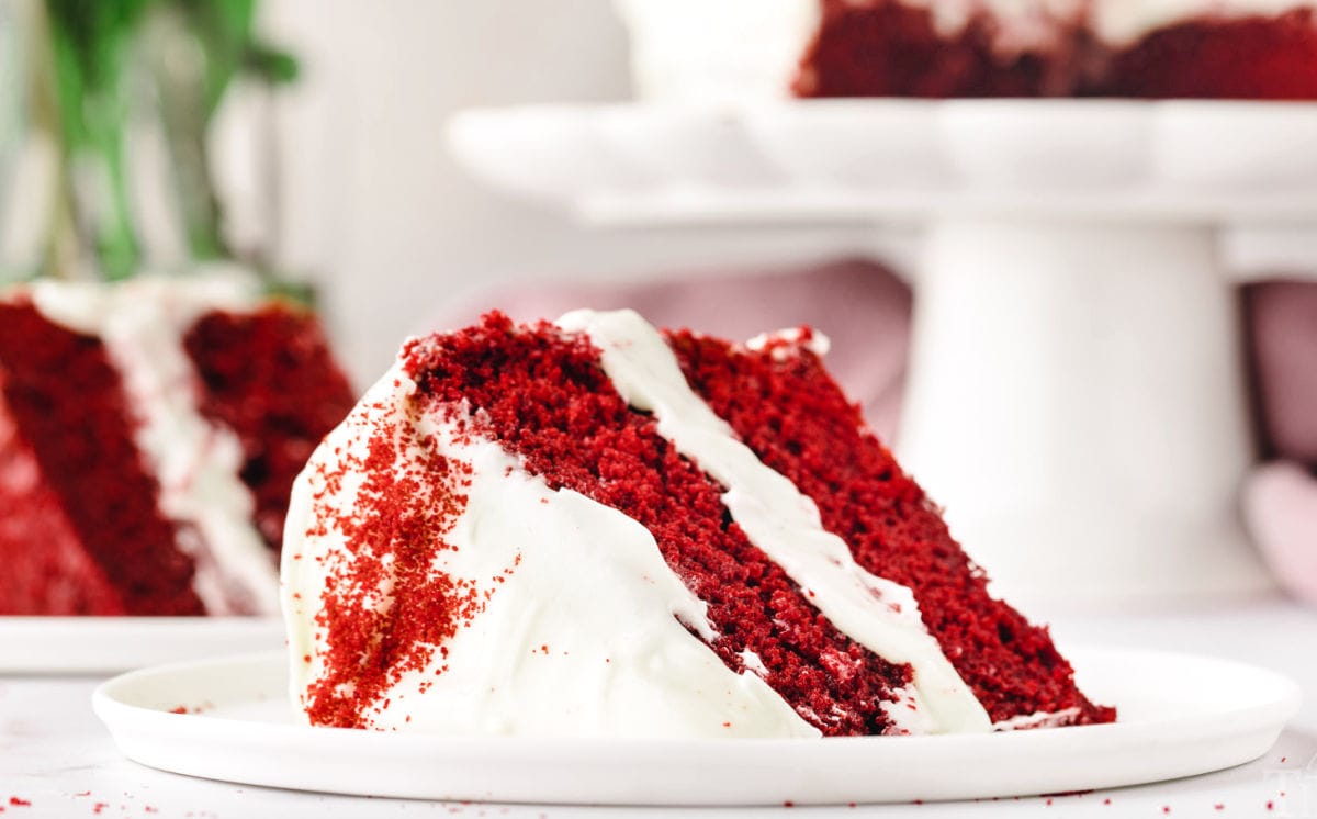 slice of red velvet cake on a white plate. another slice can be seen in the background.