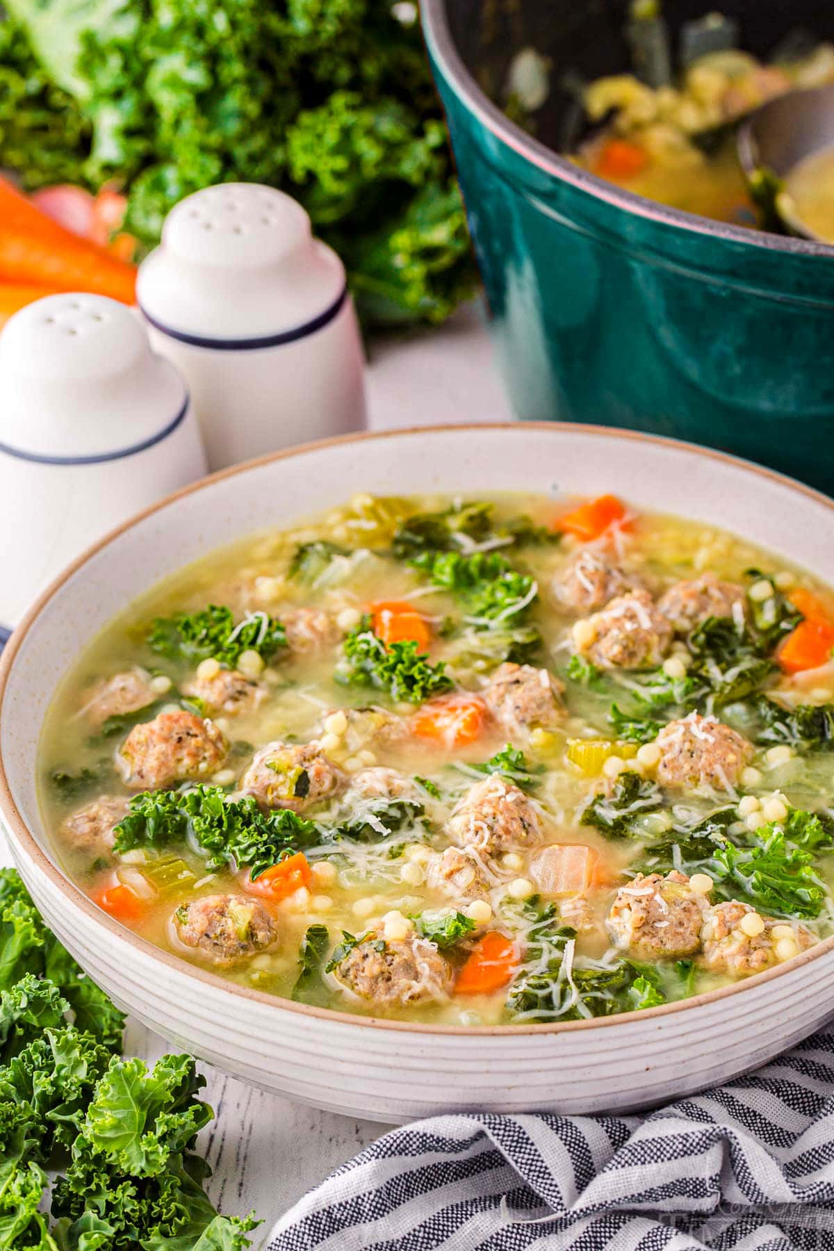 large bowl filled with Italian Wedding Soup and garnished with Parmesan cheese. A dark teal dutch oven can be seen in the background. A black and white striped towel is in the front of the bowl.