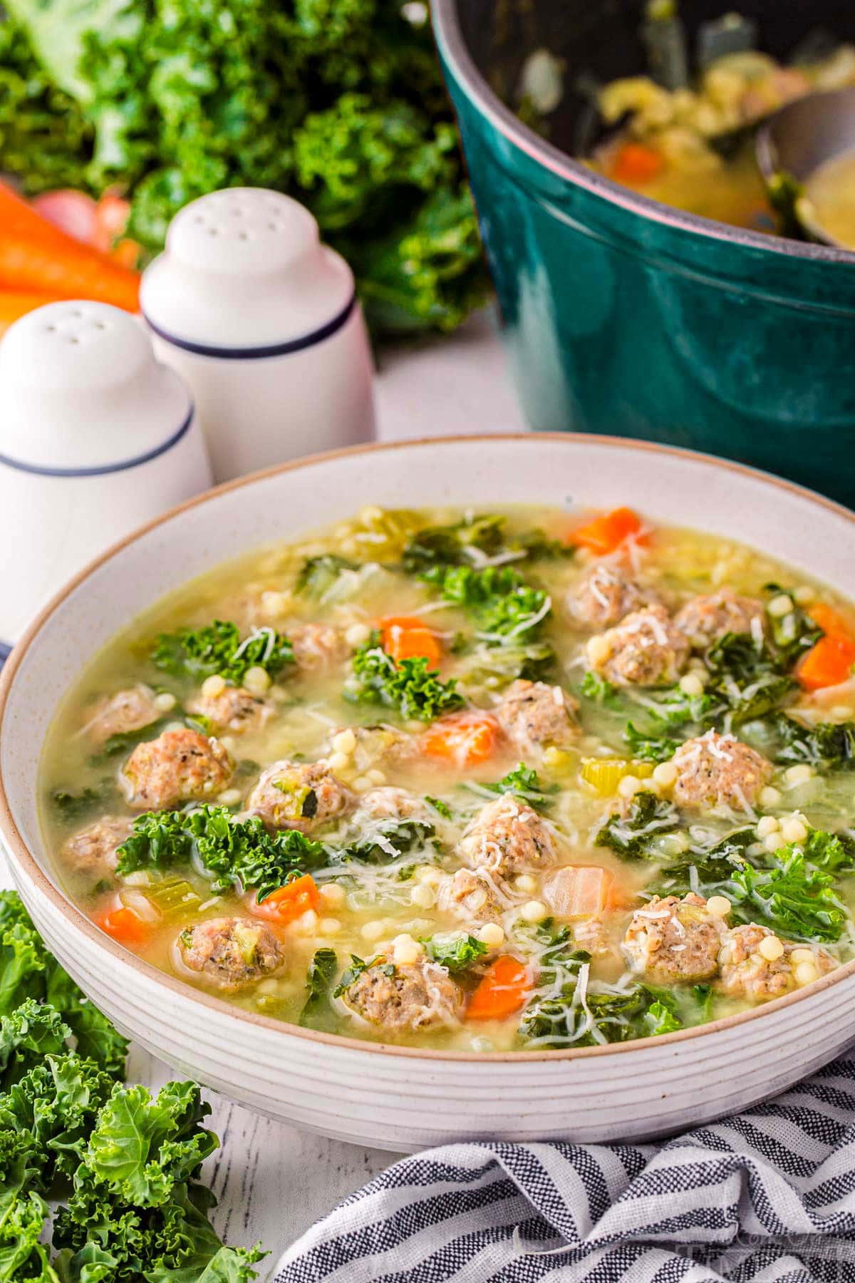 large bowl filled with Italian Wedding Soup and garnished with Parmesan cheese. A dark teal dutch oven can be seen in the background. A black and white striped towel is in the front of the bowl. 