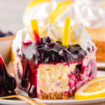 slice of lemon blueberry cheesecake topped with blueberry pie filling, whipped cream and two lemon slices sitting on small round plate with one bite taken from the top if the slice.