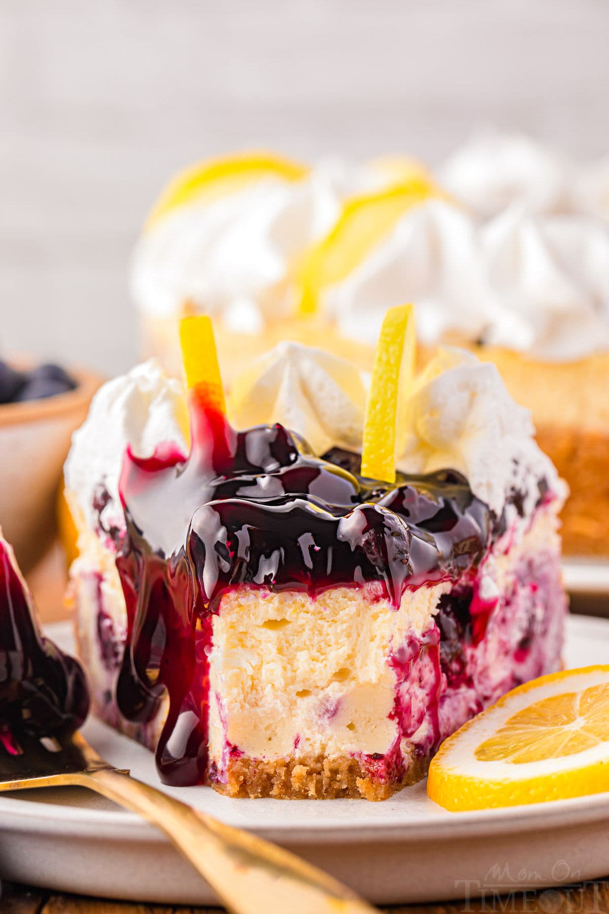 slice of lemon blueberry cheesecake topped with blueberry pie filling, whipped cream and two lemon slices sitting on small round plate with one bite taken from the top if the slice.