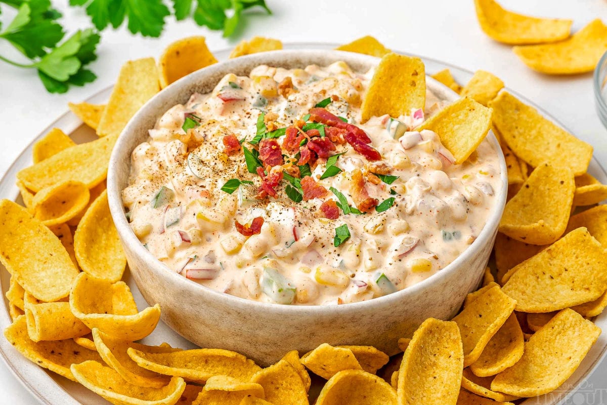 big bowl of corn dip surrounded by fritos scoops on a platter. the dip is topped with parsley and bacon bits.