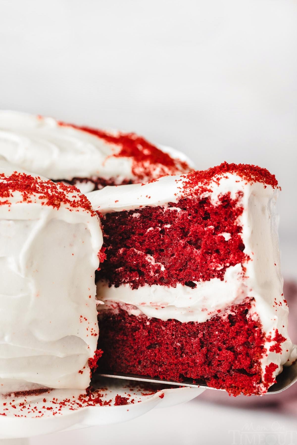 slice of red velvet cake being pulled out from the rest of the whole cake on a white cake stand.