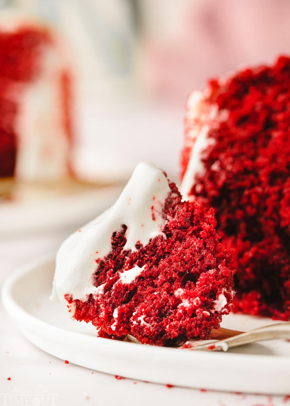 bite of red velvet cake with cream cheese frosting on gold fork on a white plate.