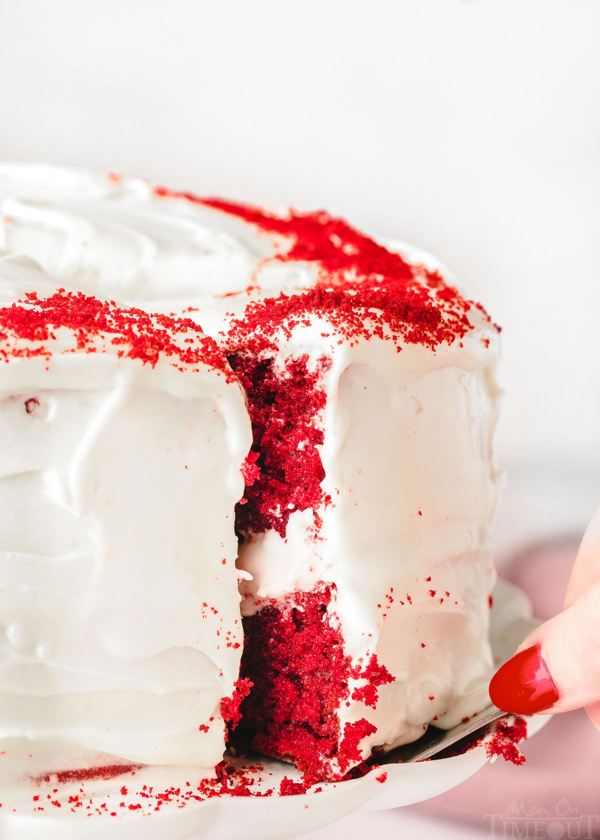 slice of red velvet cake being pulled out from the rest of the whole cake on a white cake stand.