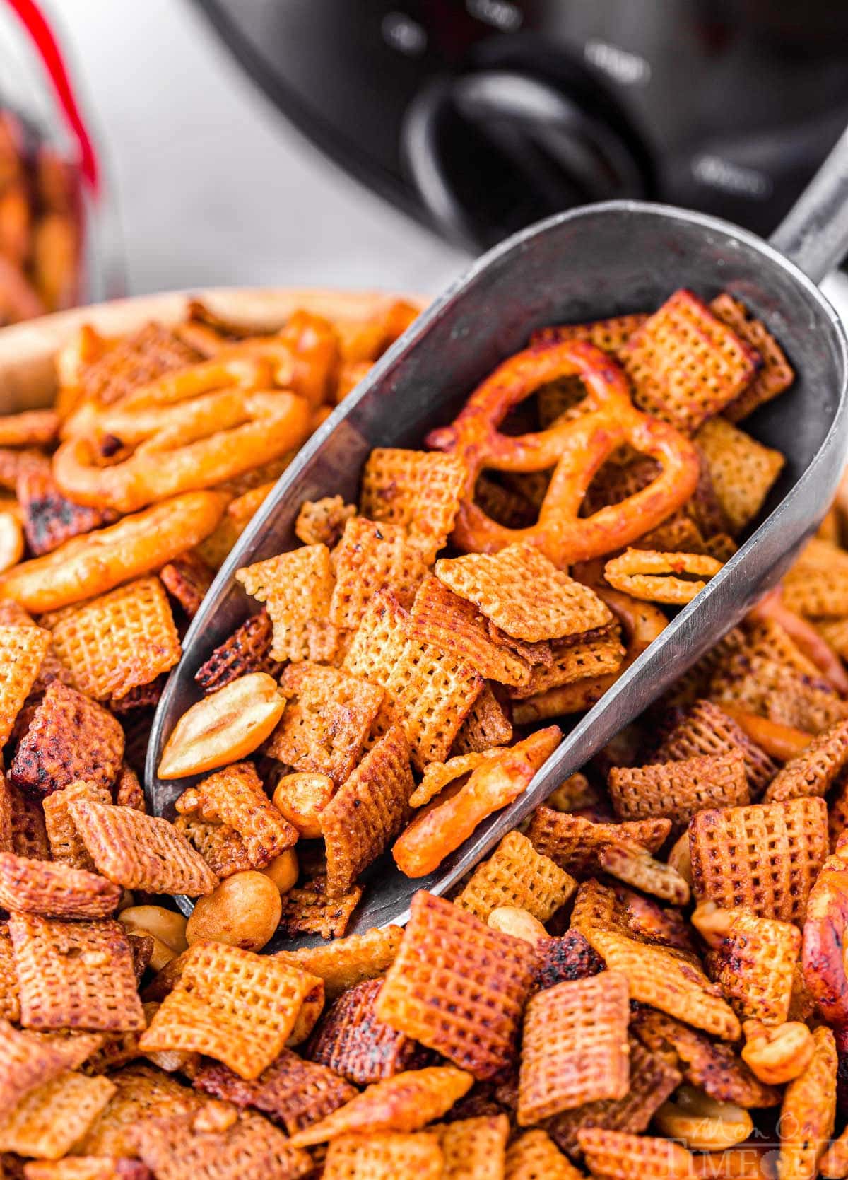 large wood bowl filled with chex mix and a large metal scoop is scooping some out. the crockpot can be seen in the background.