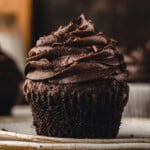 chocolate cupcake with liner removed sitting on white plate. cupcake has been topped with a chocolate buttercream frosting.