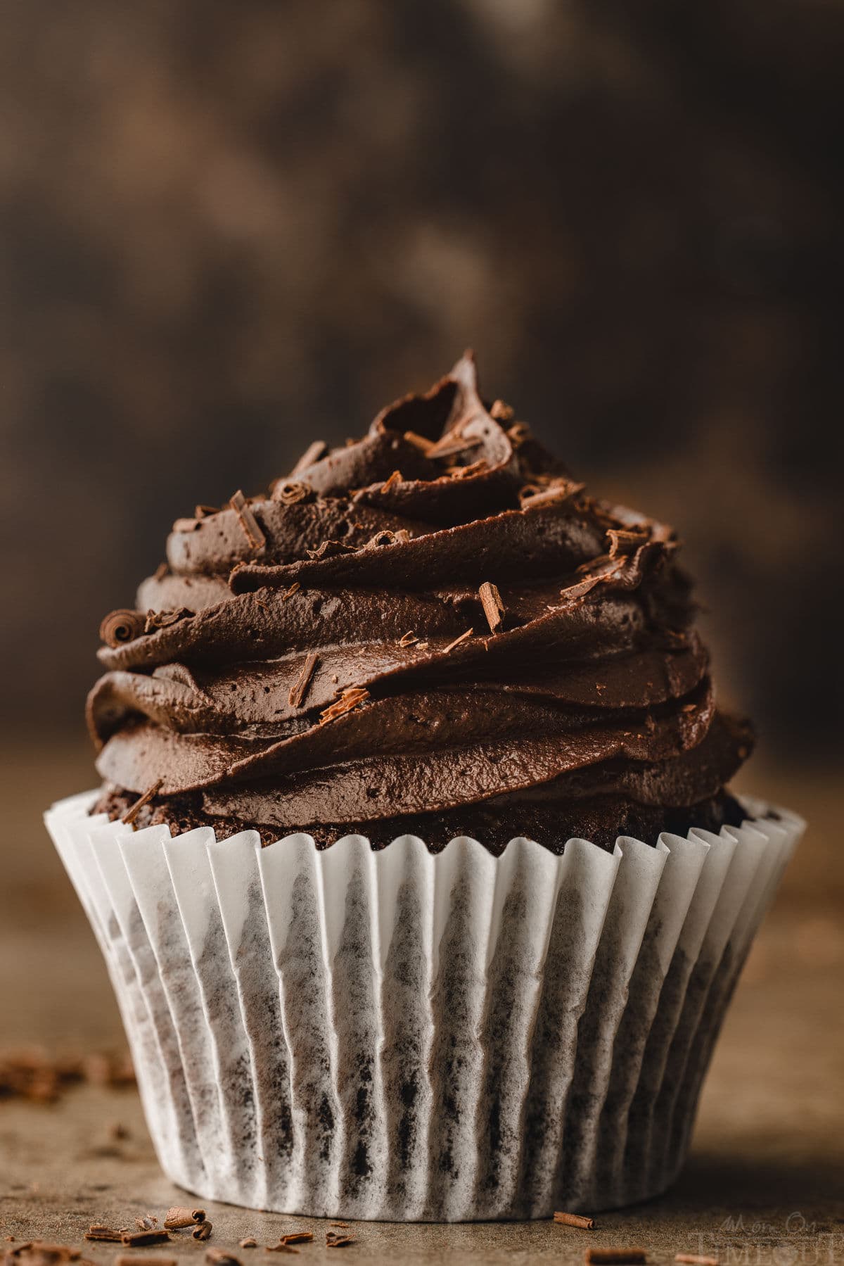 perfect chocolate cupcake topped with a deep chocolate buttercream. the cupcake is still in it's white wrapper. the background is dark and moody. 