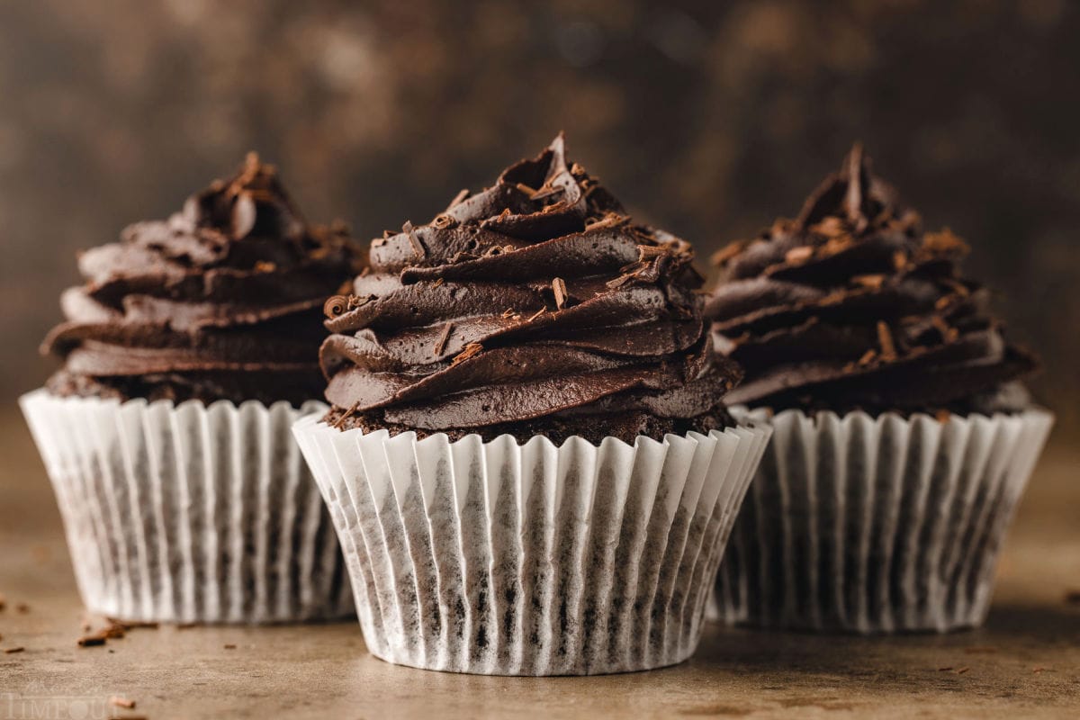 three chocolate cupcakes lined up in a row with white parchment paper liners topped with piped chocolate buttercream.